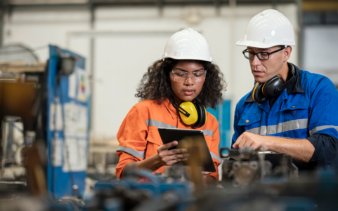 Two workers in hard hats and PPE review a tablet together next to industrial machinery.