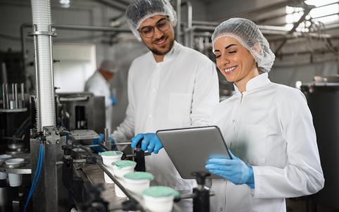 Two employees in white coats and hairnets reviewing a tablet at a food production line.